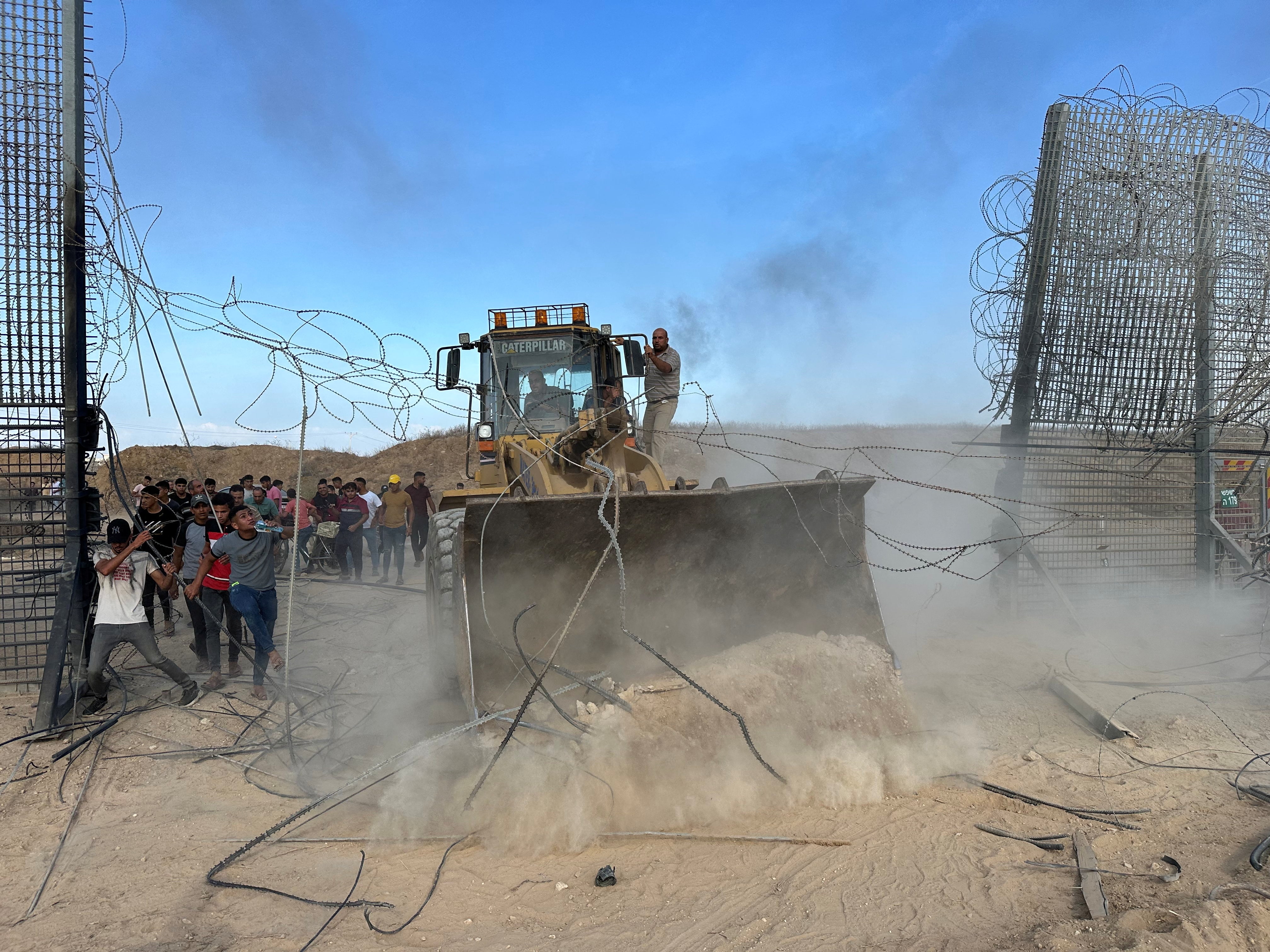 Bulldozer being driven into a tall wire mesh fence and a group of about 30 men clustered next to it.