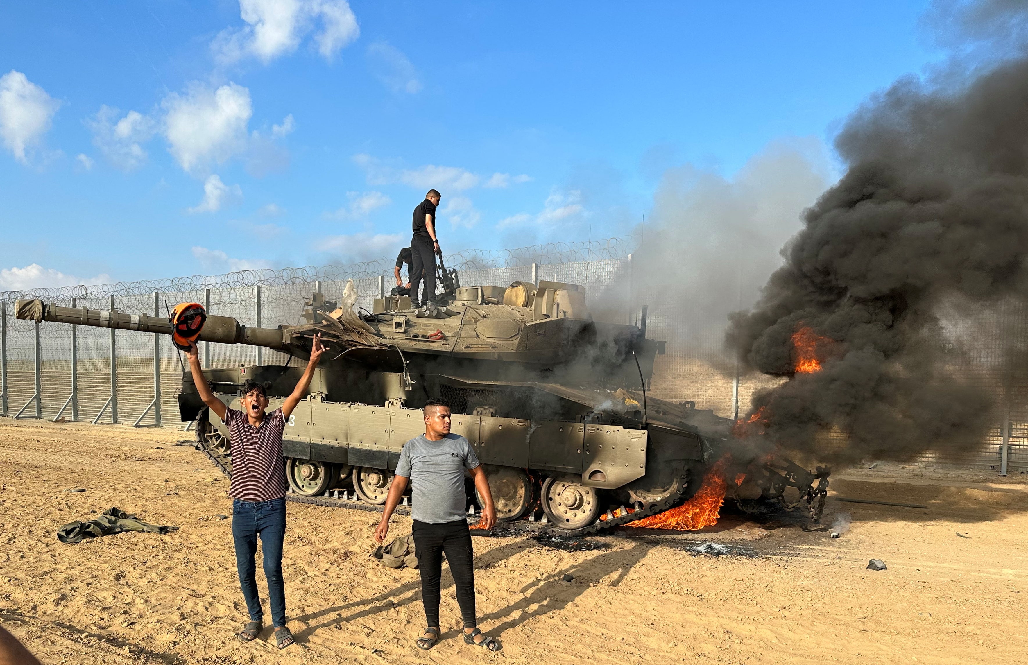 A burning military vehicle, with two men standing on top of it and two men in front.