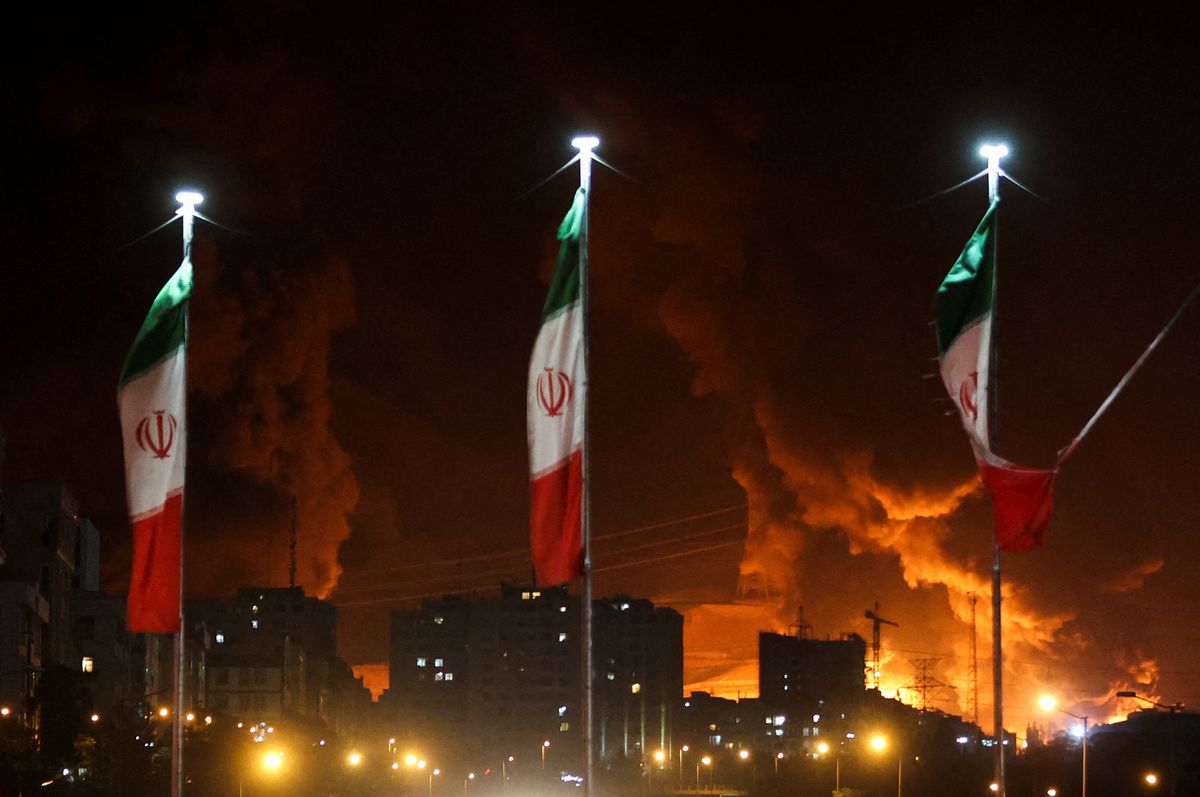 Three Iranian flags against a backdrop of silhouetted buildings and fire.